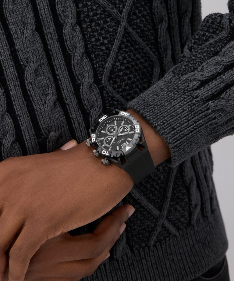 Person wearing a black knit sweater and dark gray gloves with a GUESS Mens Black Multi-function Watch on a neutral background