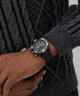 Person wearing a black knit sweater and dark gray gloves with a GUESS Mens Black Multi-function Watch on a neutral background