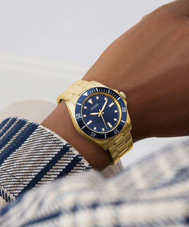 Gold GUESS Mens Gold Tone Analog Watch with a blue dial on a wrist, worn by a person with a plaid garment.
