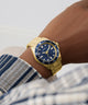 Gold GUESS Mens Gold Tone Analog Watch with a blue dial on a wrist, worn by a person with a plaid garment.