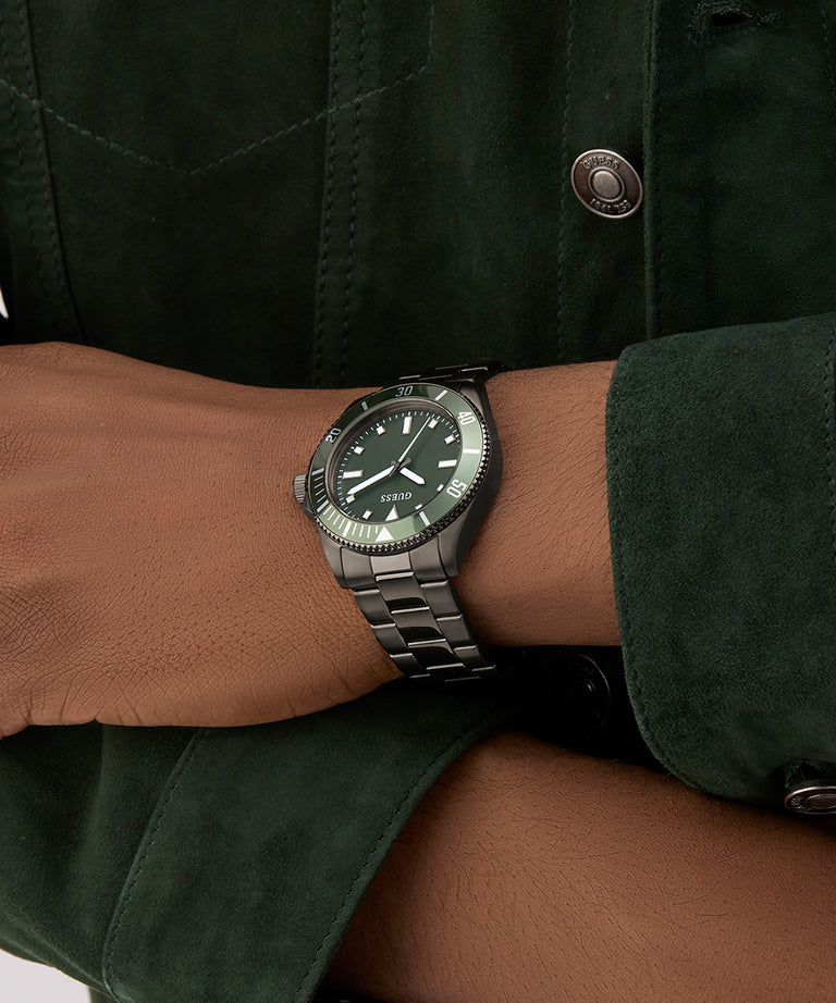 Person wearing a GUESS Mens Gunmetal Analog Watch on a wrist with a green jacket.