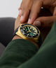 Close-up of a wrist wearing a GUESS Mens Black Gold Tone Analog Watch watch with a green face.