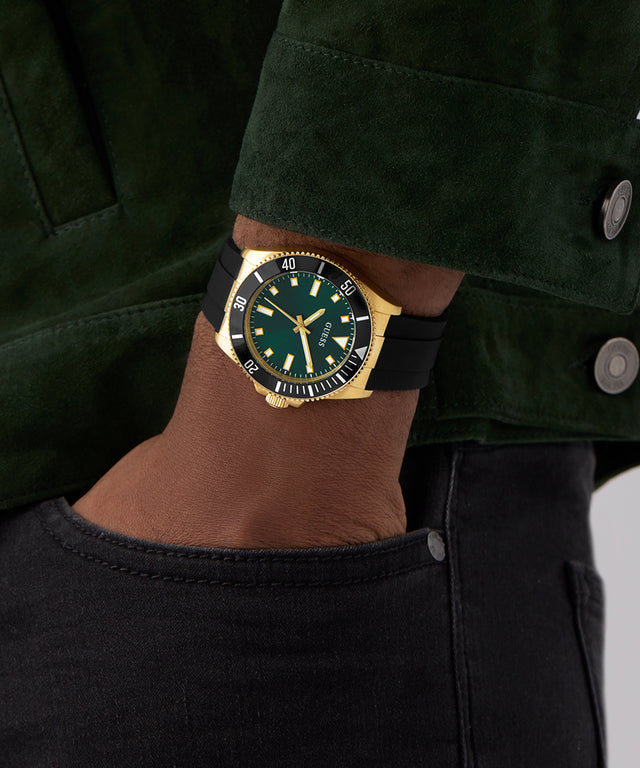 Person wearing a GUESS Mens Black Gold Tone Analog Watch with a green face and gold accents on a dark background