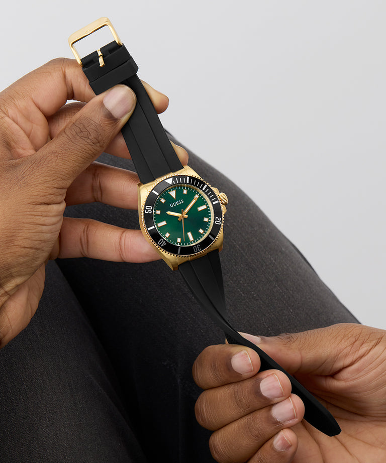 Person holding a GUESS Mens Black Gold Tone Analog Watch with a black strap and green face against a neutral background