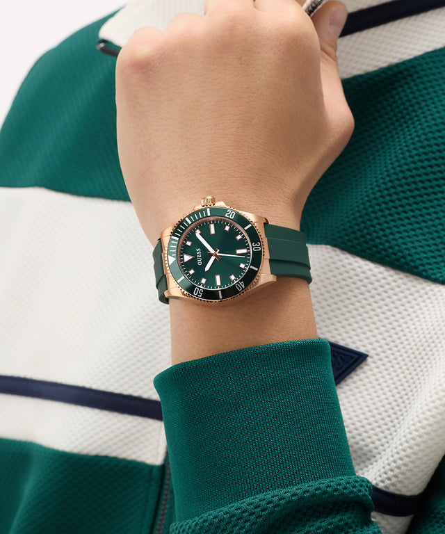 Hand wearing a green GUESS Mens Green Rose Gold Tone Analog Watch with a green strap against a green and white striped background