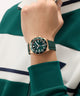 Hand wearing a green GUESS Mens Green Rose Gold Tone Analog Watch with a green strap against a green and white striped background