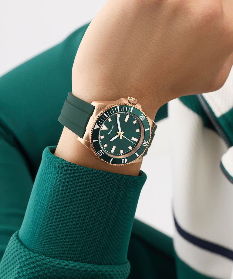 Person wearing a green GUESS Mens Green Rose Gold Tone Analog Watch with a rose gold case on a blurred background