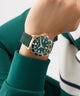 Person wearing a green GUESS Mens Green Rose Gold Tone Analog Watch with a rose gold case on a blurred background