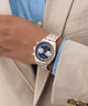 Close-up of a person wearing a silver and gold GUESS Mens 2-Tone Multi-function Watch with a blue face, dressed in a beige suit and blue shirt.