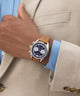 Hand wearing a watch with a brown leather strap and blue face, against a beige suit background.