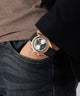 Person wearing a gold GUESS Mens Black Rose Gold Tone Multi-function Watch with a black strap on a neutral background