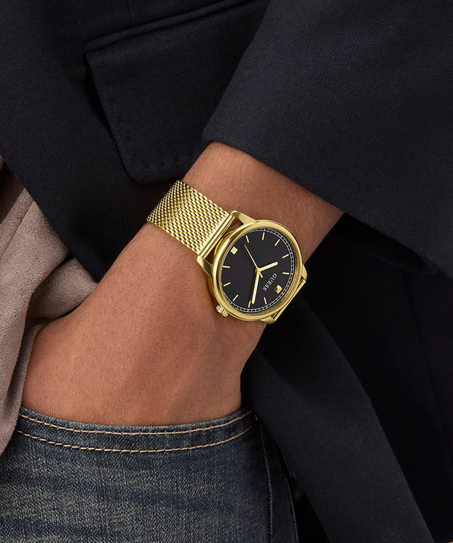 GUESS Mens Gold Tone Analog Watch on a wrist with a black face, worn by a person in a dark suit and jeans.