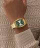 Gold wristwatch with a green face on a wrist wearing a beige sweater. GW0975G2