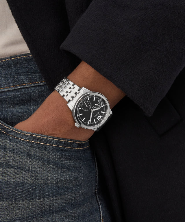 Silver GUESS Mens Silver Tone Multi-function Watch on a person's wrist with a black coat and jeans in the background