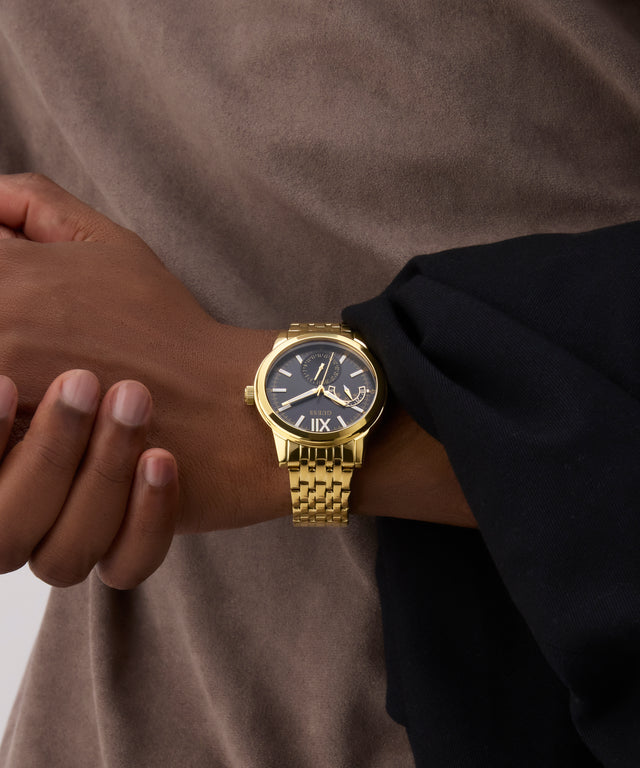Gold GUESS Mens Gold Tone Multi-function Watch on a person's wrist with a neutral background