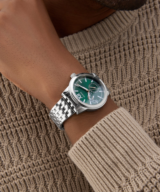 Green GUESS Mens Silver Tone Multi-function Watch with silver bracelet on a wrist against a textured brown background