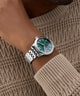 Green GUESS Mens Silver Tone Multi-function Watch with silver bracelet on a wrist against a textured brown background