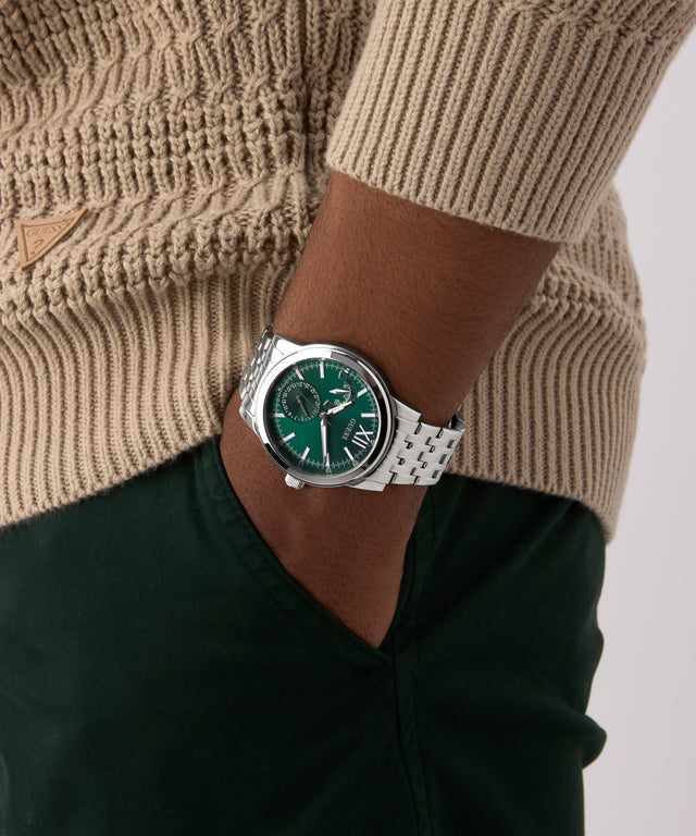 Person wearing a green GUESS Mens Silver Tone Multi-function Watch with a silver strap on a neutral background