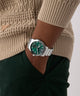 Person wearing a green GUESS Mens Silver Tone Multi-function Watch with a silver strap on a neutral background