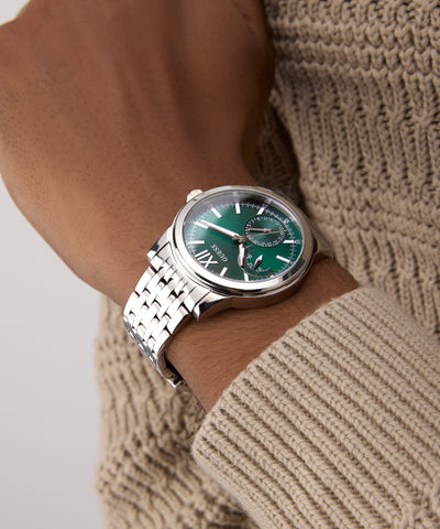 Close-up of a wrist wearing a silver watch with a green face, against a neutral background.