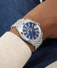 GUESS Mens Silver Tone Multi-function Date Watch with blue dial on a wrist, wearing a white sleeve and dark jeans.