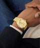 Gold GUESS Mens Gold Tone Multi-function Date Watch on a person's wrist with a blurred background