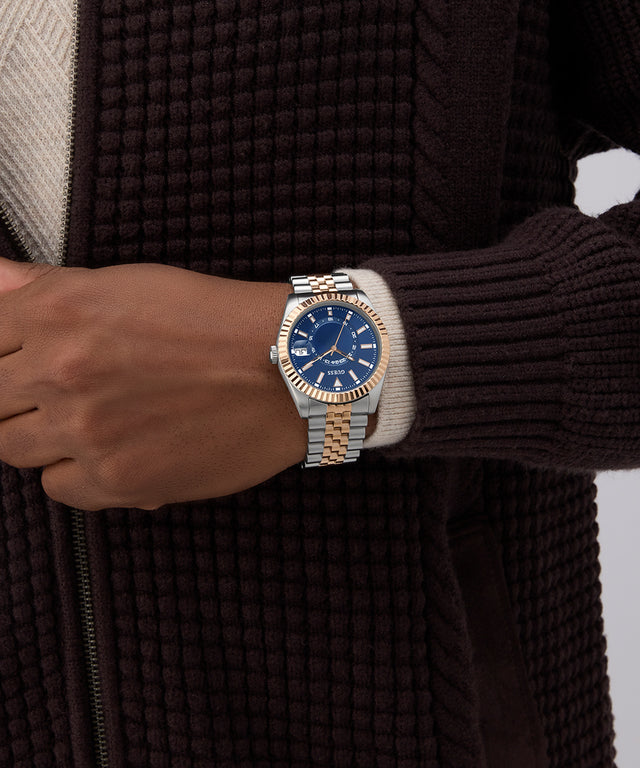 Hand wearing a two-tone watch with a blue face, against a dark textured background. GW0977G5