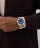 Hand wearing a two-tone watch with a blue face, against a dark textured background. GW0977G5