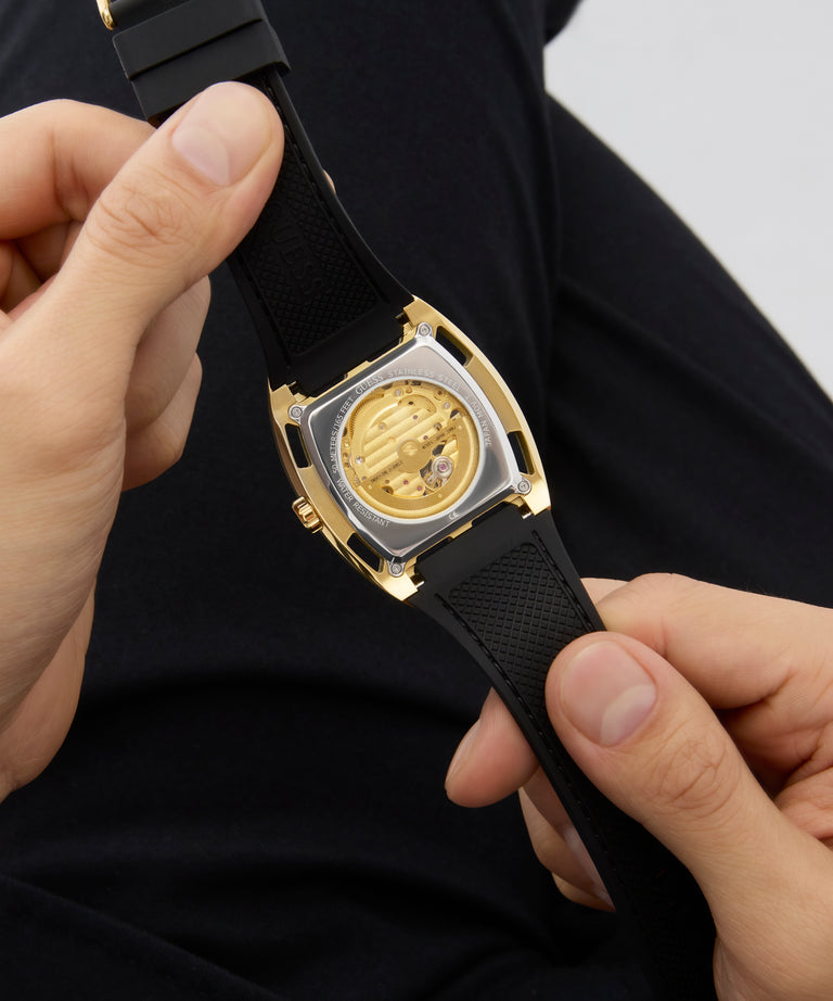 GUESS Mens Black Gold Tone Automatic Watch caseback held by a person against a dark background