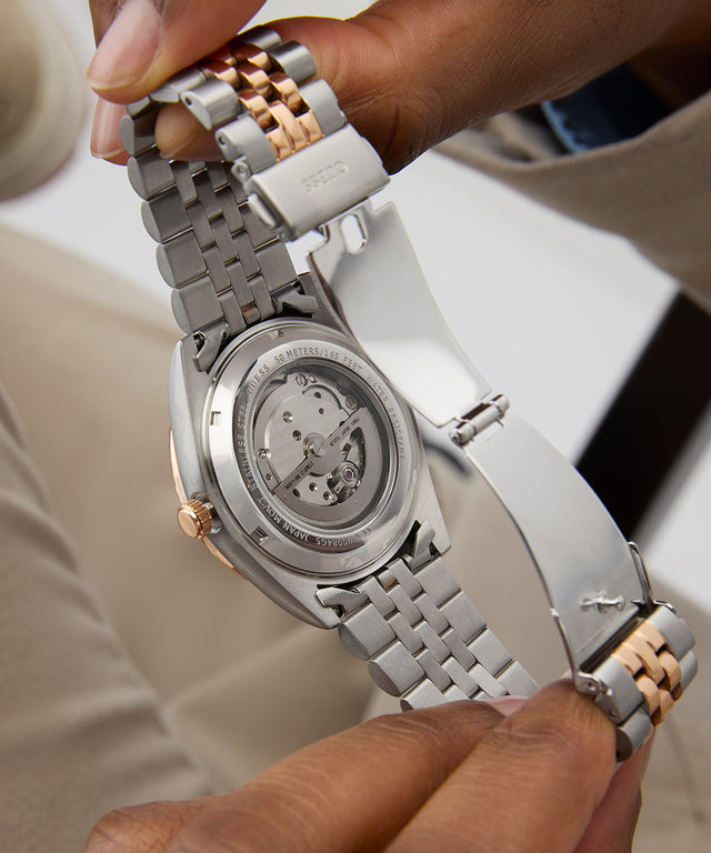 GUESS Mens 2-Tone Automatic Watch with a detailed mechanism held by a hand against a neutral background