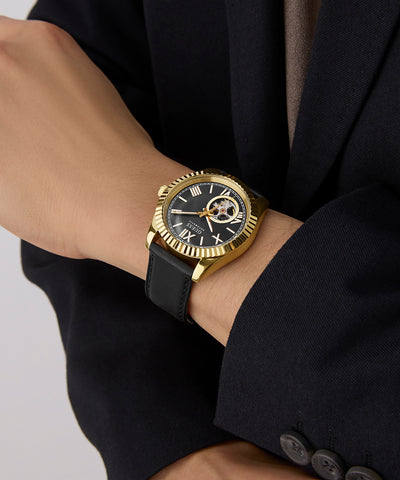 GUESS Mens Black Gold Tone Automatic Watch with black strap on a person's wrist wearing a black suit jacket.