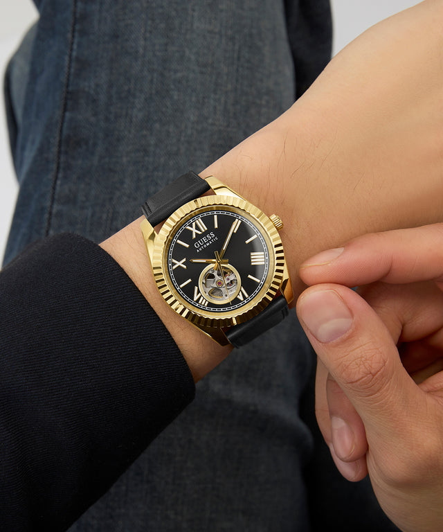 GUESS Mens Black Gold Tone Automatic Watch with black strap on a wrist against a neutral background