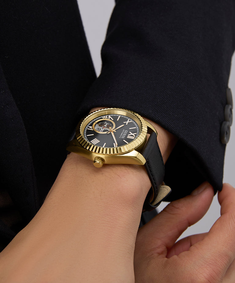GUESS Mens Black Gold Tone Automatic Watch with black strap on a wrist against a dark background
