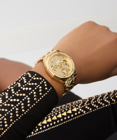 GUESS Ladies Gold Tone Multi-function Watch on a wrist with a black and gold patterned sleeve against a light gray background