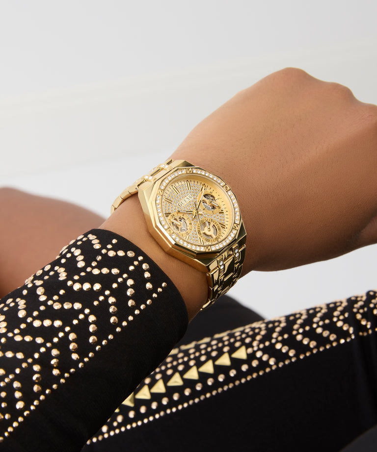 GUESS Ladies Gold Tone Multi-function Watch on a wrist with a black and gold patterned sleeve against a light gray background
