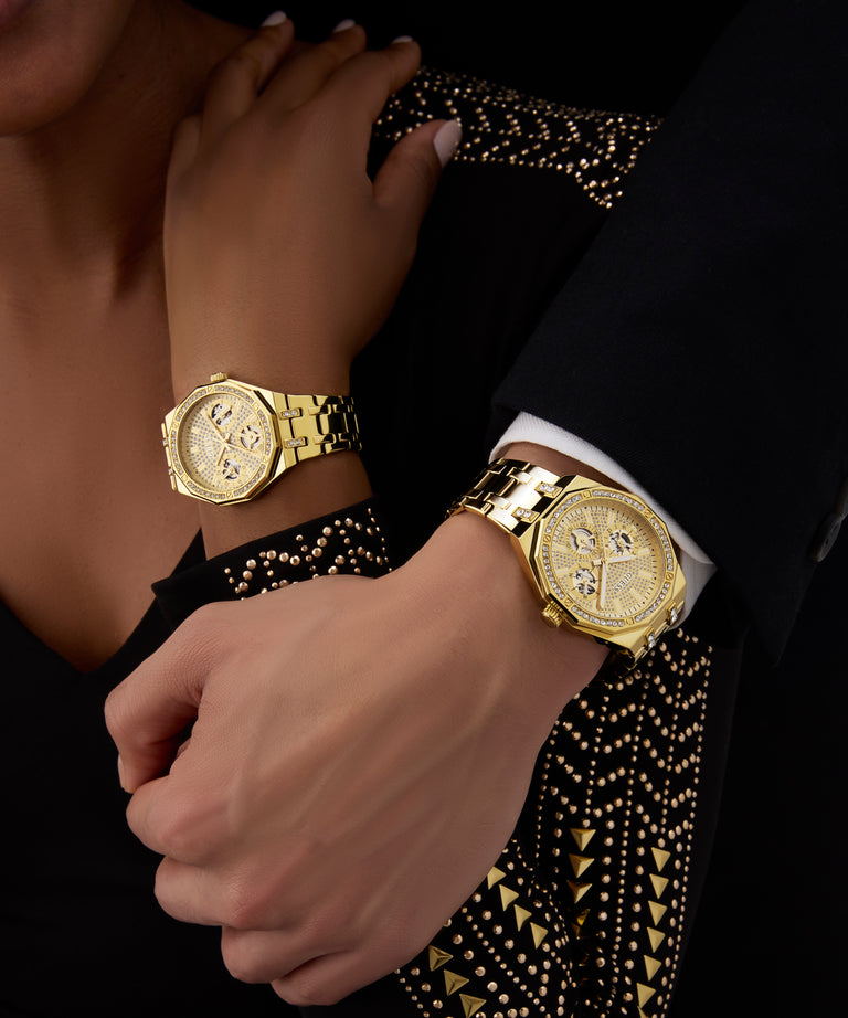 Close-up of two hands wearing GUESS Ladies Gold Tone Multi-function Watch GUESS Ladies Gold Tone Multi-function Watch on a black and gold patterned background