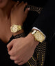 Close-up of two hands wearing GUESS Ladies Gold Tone Multi-function Watch GUESS Ladies Gold Tone Multi-function Watch on a black and gold patterned background