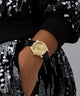 Gold GUESS Ladies Black Gold Tone Analog Watch on a wrist with a black strap against a sequined background