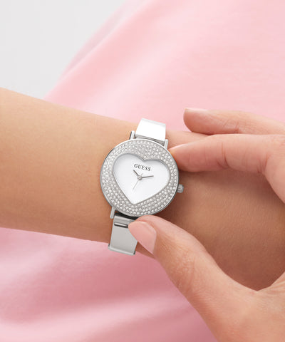 Silver heart-shaped GUESS Ladies Silver Tone Analog Watch with gemstones on a wrist against a pink background