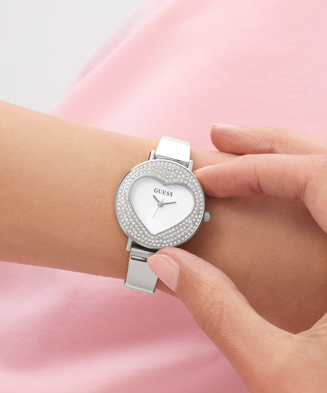 Silver heart-shaped GUESS Ladies Silver Tone Analog Watch with gemstones on a wrist against a pink background