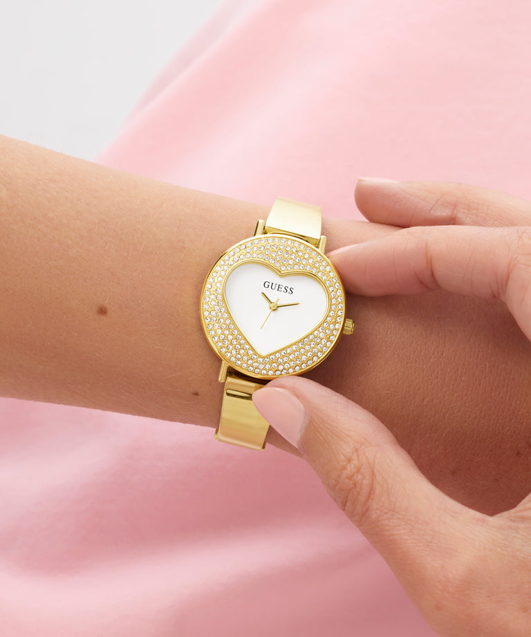 Gold heart-shaped GUESS Ladies Gold Tone Analog Watch with gemstones on a wrist against a pink background