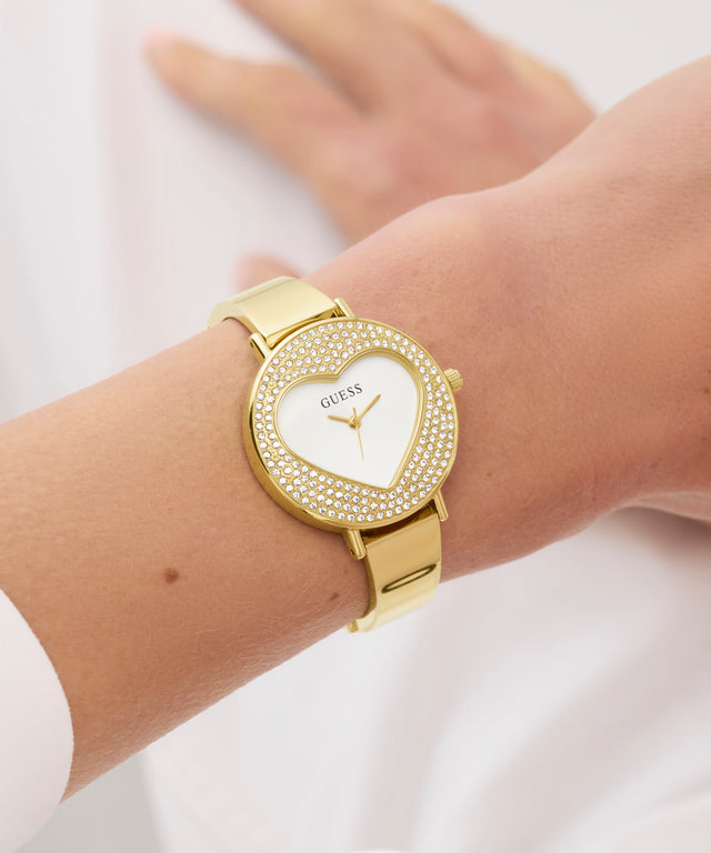 GUESS Ladies Gold Tone Analog Watch with heart-shaped face on a wrist against a white background