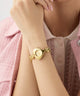 GUESS Ladies Valerie Gold Tone Analog Watch on a person's wrist with a soft pink background