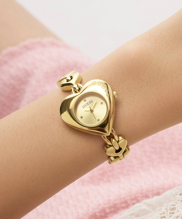 Gold heart-shaped GUESS Ladies Valerie Gold Tone Analog Watch on a wrist with a soft pink background
