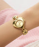 Gold heart-shaped GUESS Ladies Valerie Gold Tone Analog Watch on a wrist with a soft pink background