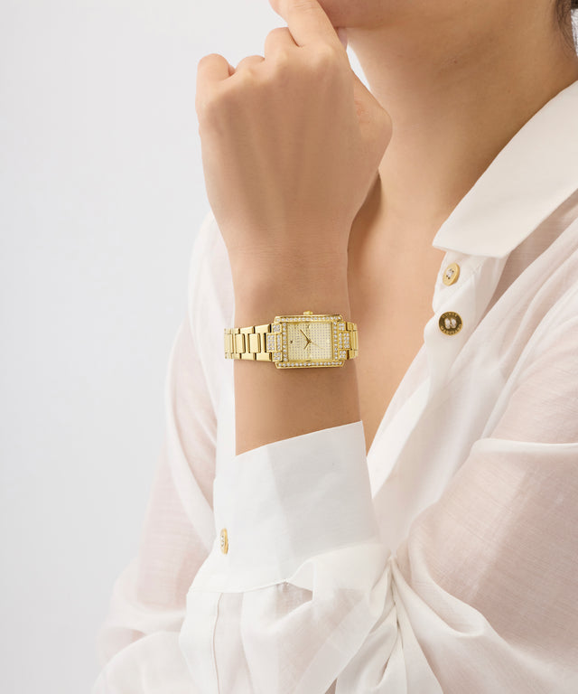 Gold GUESS Ladies Gold Tone Analog Watch on a wrist with a white shirt against a light background