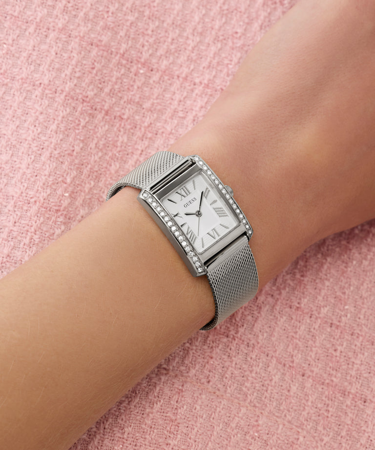 Silver GUESS Ladies Silver Tone Analog Watch with mesh band on a wrist against a pink textured background