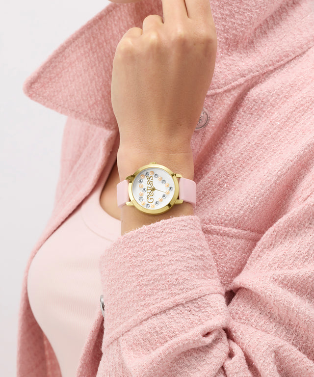 Hand wearing a GUESS Ladies Pink Gold Tone Analog Watch with a pink strap against a pink background
