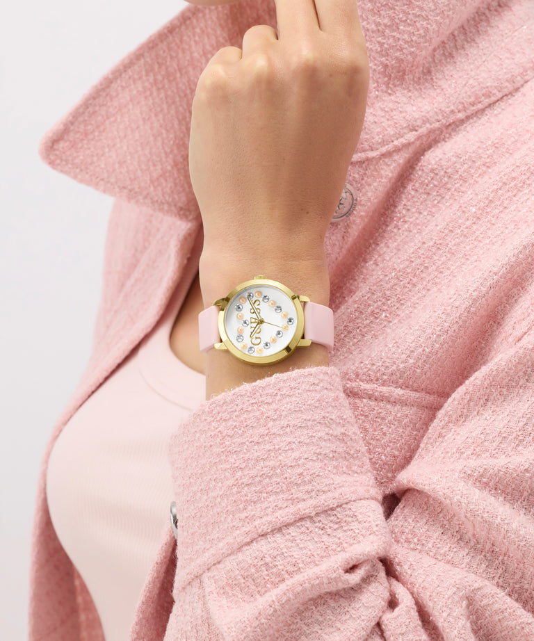 Hand wearing a GUESS Ladies Pink Gold Tone Analog Watch with a pink strap against a pink background