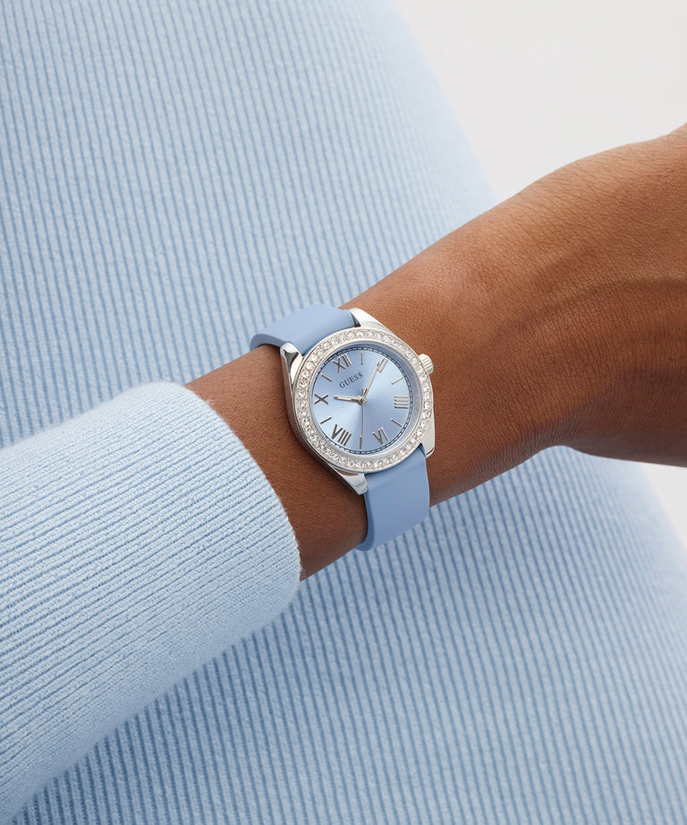 Wrist wearing a light blue GUESS Ladies Mini Luna Blue Silver Tone Analog Watch with a silver case against a light blue background